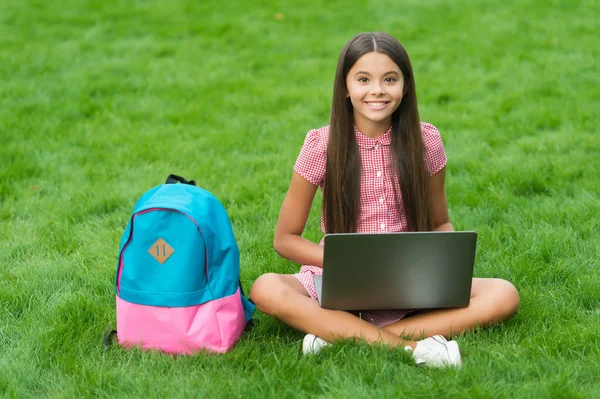 Ragazza reagisce durante l'utilizzo di un computer portatile sedersi su erba verde nel parco con la schiena della scuola, l'istruzione — Foto Stock