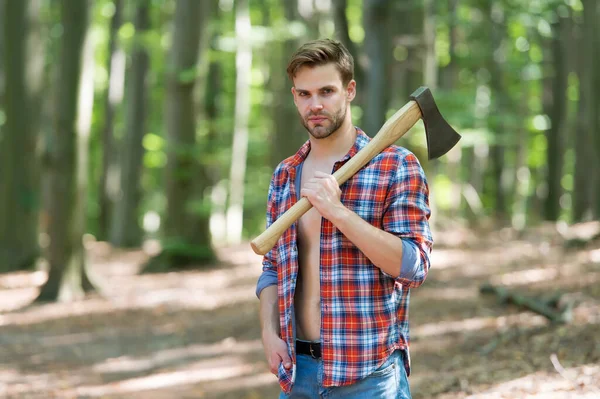 Unshaven handsome man in open plaid shirt with jeans carry large splitting axe in summer forest natural landscape, lumberman