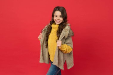 Uzun kıvırcık saçlı gülümseyen çocuk kırmızı arka plan, moda ve güzellikte kalın örgü süveter ve kürk yelek giyer.