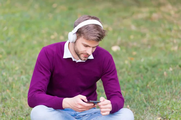 녹색 잔디에 있는 잘생긴 남자 휴대 전화 mp3 플레이어에서 노래를 바꾸고 디지탈 시대의 이어폰으로 음악을 듣습니다. — 스톡 사진