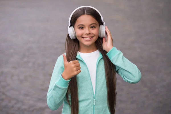 Happy smiling child enjoys listens to music in headphones. kid listen song outdoor. walking with favorite tune. education online concept. childhood happiness. elearning of student girl. thumb up — Stock Photo, Image