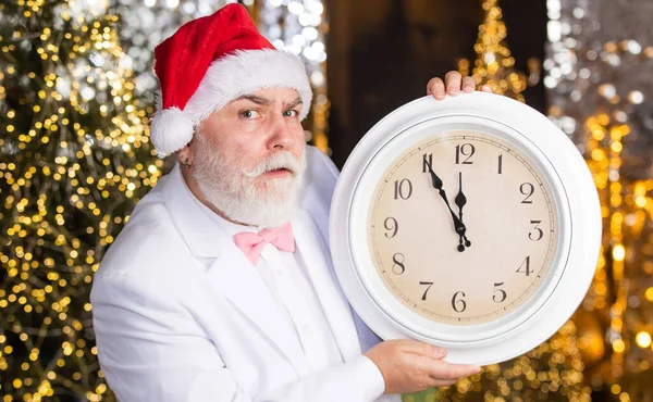 Štědrý večer. Santa Claus počítá čas. Půlnoční koncept. Nový rok odpočítávání. Muž s retro hodinami. Pozadí osvícení věnce. Vousatý obchodník v Santově klobouku. Senior Santa Man ve smokingu — Stock fotografie