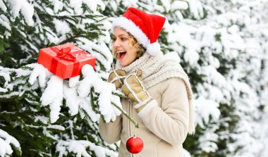 Vay canına, neşeli ol. Mutlu kadının elinde Noel hediyesi kutusu var. Noel ağacı süslemeli kız. Bayan Kırmızı Noel Baba şapkası kürk ağacında. Kış alışveriş mutluluğu. Açık hava mükemmel bir ruh halidir. satış ve indirim