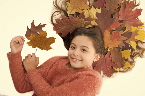 Prevent dry split hair ends. Child enjoy fall season. Girl cute kid long hair lay on white background with fallen leaves. Dry maple leaves in hairstyle. Fall season concept. Hair care in autumn