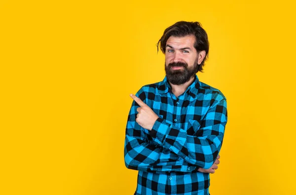 Retrato de um homem de barba sorridente em camisa quadriculada apontando os dedos para longe no espaço de cópia sobre fundo amarelo, apresentando produto — Fotografia de Stock