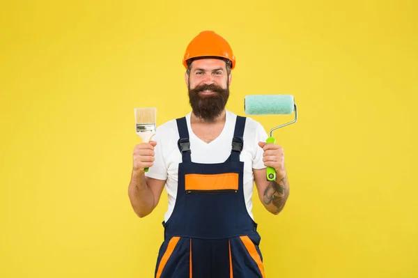 İş üniformalı mutlu fırça ressamı yüzey boyama ve sarı arkaplan dekorasyonu için araçları tutar, yeniden boyar — Stok fotoğraf