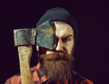 Şapkalı yakışıklı sakallı adam siyah arka planda balta tutuyor.