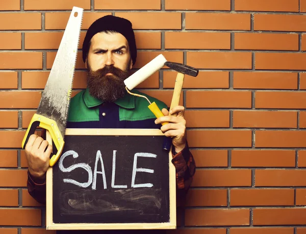 Bearded man holding various building tools and board, serious face — Stock Photo, Image