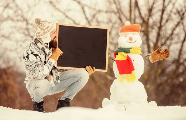 Winter holidays. Upcoming event. Man with beard hold chalkboard copy space. Winter announcement. Hipster knitted hat and gloves show blackboard. Guy and snowman snowy nature background. Winter event
