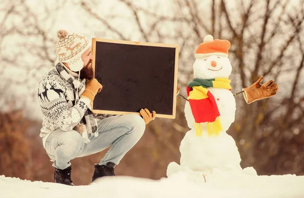 Vacaciones de invierno. Próximo evento. El hombre con barba tiene espacio para copiar pizarras. Anuncio de invierno. Hipster sombrero de punto y guantes muestran pizarra. Guy y muñeco de nieve fondo de la naturaleza. Evento de invierno — Foto de Stock