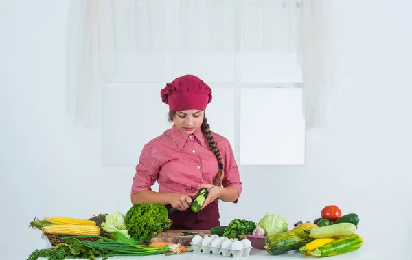 Pelar un pepino. adolescente chica preparando comida. verduras frescas crudas. Hora de comer. feliz niño cocinando en la cocina. solo alimentos saludables y orgánicos. Lleno de vitaminas. niño llevar uniforme de chef y sombrero — Foto de Stock