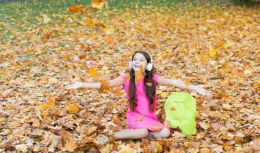 Acele etme. Modern eğitim. Sonbahar ormanında okul çantalı bir çocuk. Bilgi günü konsepti. Sonbahar parkındaki çocuk. Mutlu çocuk sonbahar yapraklarıyla eğleniyor. Küçük kız kulaklık takıyor. Okula geri dön.