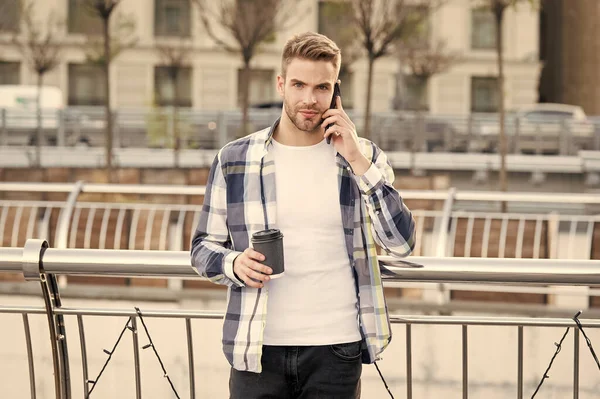 Obchodník ve městě. student s kávou pomocí mobilního telefonu. Mladý muž pije kávu na ulici a používá mobilní telefon. samec používající chytrý telefon v moderním životě. muž mluví po telefonu držet šálek kávy — Stock fotografie