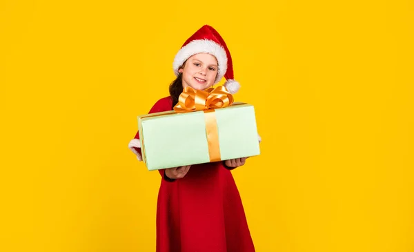 Kislány Mikulás kalap tartó ajándék doboz. A gyerek tartsa a jelenlegi doboz sárga háttér. Boldog karácsonyt és boldog ünnepeket! Az álmok valóra válnak. Téli ünnepek. Az én boldog napom. Karácsonyi ajándékvásárlás. Ajándékcsomag — Stock Fotó