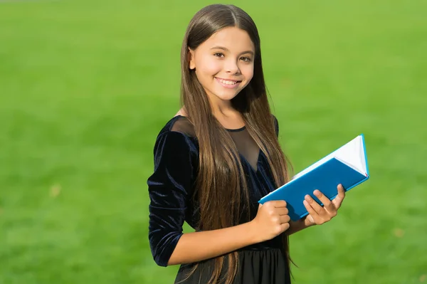 Glad liten unge i skolan uniform leende hålla studie bok grönt gräs solig sommar utomhus, utbildning — Stockfoto