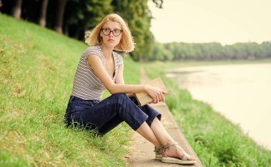 Dinlen ve hobi olarak dinlen. Tatil sezonu. Yaz tatili. Kız nehir kenarındaki yeşil çimlerde oturuyor. Kız iş gününden sonra nehir kenarında dinleniyor. Tatil hayali kuran kadın. Kendine zaman ayır.