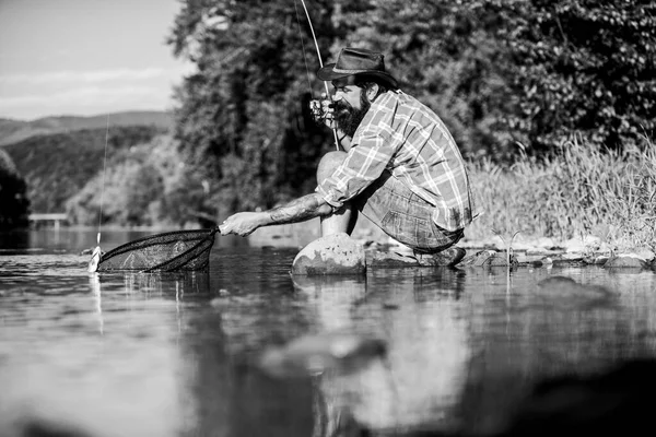 Pescador exitoso en el agua del lago. pesca de caza mayor. relajarse en la naturaleza. pesca hipster con cuchara-cebo. hombre barbudo maduro con pescado en caña. pasatiempo peces mosca. Actividad de verano. Qué pez. — Foto de Stock