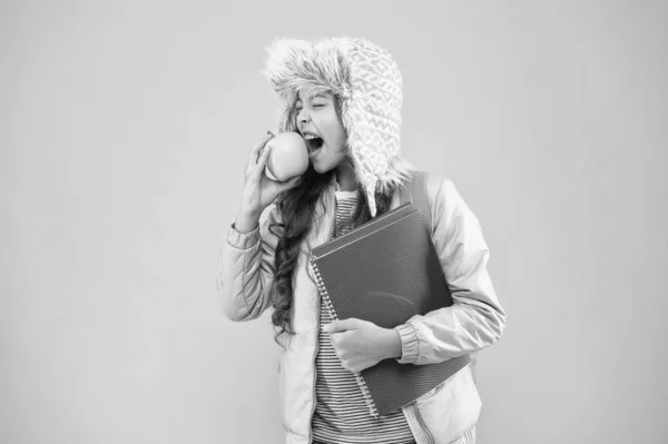 Školní prázdniny. malá šťastná dívka ušní klapka klobouk jíst jablko. zimní dovolená a dovolená. dospívající dítě růžové pozadí. každodenního života žáků. Moderní vzdělání. školačka s knihami po lekci — Stock fotografie
