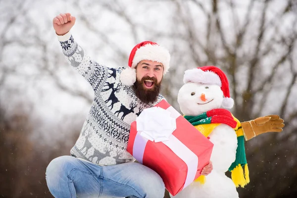 Winter games. Winter activity. Winter vacation. Man made snowman. Man Santa hat having fun outdoors. Guy happy face snowy nature background. Hipster with beard hold gift box. Surprise concept