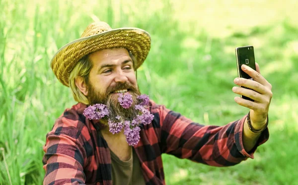 Homem maduro jardineiro relaxar na grama verde. desfrutar da natureza primavera. selfie verão com lilás no telefone. rancho brutal hipster tem longa barba saudável. flor no cabelo. agricultor relaxado em chapéu de palha — Fotografia de Stock