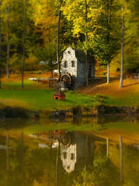 Tilt shift fotoğraf eski bir taş su değirmeni yansıtılır berrak bir göl karşı bir sonbahar ormanı arkaplan.