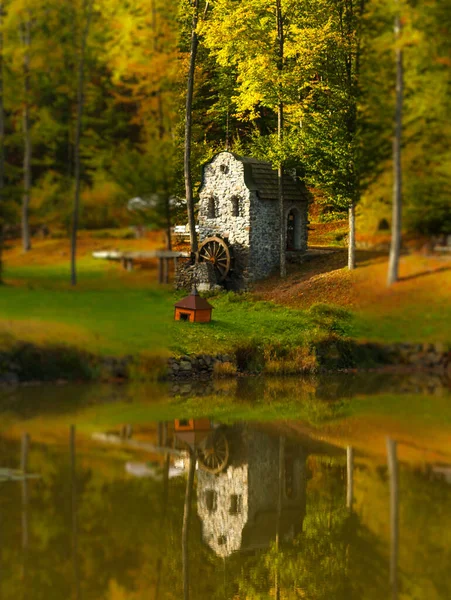 Tilt shift photograph An old stone water mill is reflected in a clear lake against the backdrop of an autumn forest.