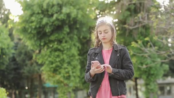 Muchacha atractiva divertida joven usando teléfono inteligente y sonrisas en el fondo de árboles verdes en verano. Chica bonita en un suéter rosa escribe un mensaje en el teléfono en medio del parque . Imágenes de stock libres de derechos