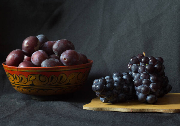 Деревянная чаша с черными сливами и черным маленьким плотным виноградом на деревянной доске. Натюрморт
.