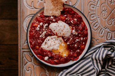 Sabah kahvaltısı. Bir tabakta iki haşlanmış yumurta ile Domates shakshuka. Yanında çıtır ekşi hamur ekmeği. Fransız basınında kahve.