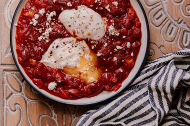 Sabah kahvaltısı. Bir tabakta iki haşlanmış yumurta ile Domates shakshuka. Yanında çıtır ekşi hamur ekmeği. Fransız basınında kahve.