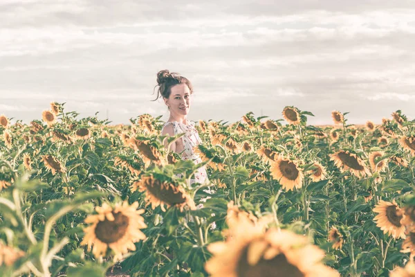 Bela senhora está andando no campo de girassol durante o pôr do sol em um vestido de verão . — Fotografia de Stock