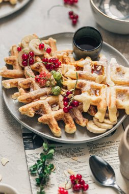 Kırmızı üzümlü çıtır Belçika waffleları, bektaşi üzümü, akçaağaç şurubu ve gri seramik tabakta pudra şekeri.