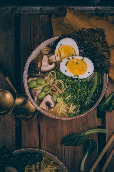 Ramen Japanese soup. Traditional Japanese cuisine. Asian broth with noodles, bok choy, shiitake mushrooms, edamame. Rustic dark mood. Boiled egg in soup. Dashi broth. Chopsticks, spoons on wood table.