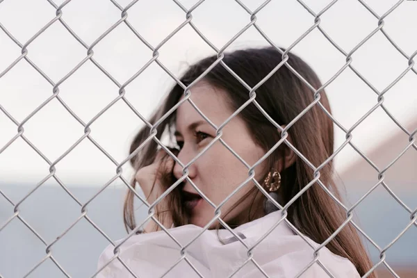 Pretty woman stands against wired fence. Model in white shirt. Purple lips. Pretty make up. Posing for magazine. Street fashion. Urban lifestyle. lady with long hair, beautiful eyes, behind fence.