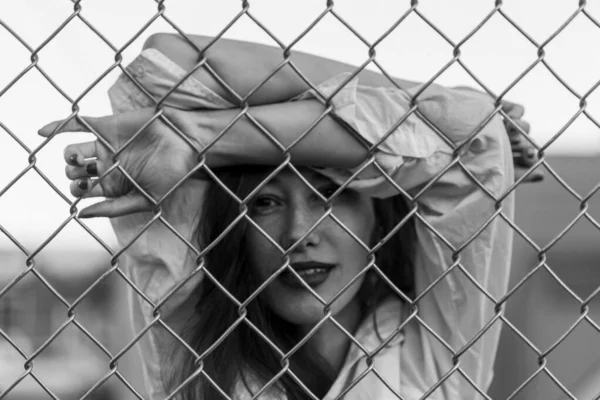 Una mujer guapa se levanta contra una valla. Modelo en camisa blanca. Labios morados. Bastante maquillaje. Posando para la revista. Moda callejera. Estilo de vida urbano. señora con el pelo largo, ojos hermosos, detrás de la cerca . —  Fotos de Stock