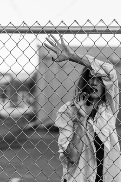 Una mujer guapa se levanta contra una valla. Modelo en camisa blanca. Labios morados. Bastante maquillaje. Posando para la revista. Moda callejera. Estilo de vida urbano. señora con el pelo largo, ojos hermosos, detrás de la cerca . —  Fotos de Stock