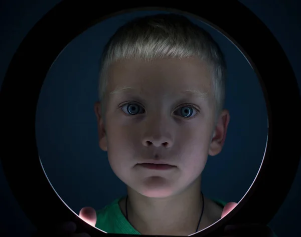 boy, blond with blue eyes. standing behind a round lamp. portrait