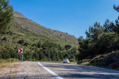 Asfalt ada yolunda araba, viraj almak üzere. Yol etrafında ağaçlar ve dağlar ile kaplı 