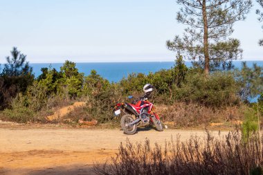 Enduro motosiklet yeşil çalılar, ağaçlar ve deniz manzarası önünde, yere park 