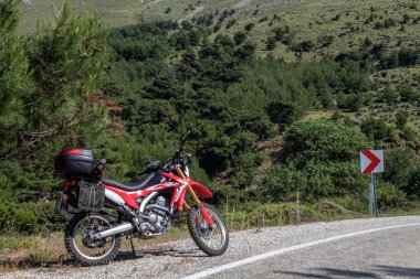   Yolun sol tarafında kırmızı bir enduro motosikleti, arka planda orman var ve asfalt zeminin üzerinde duruyor. 
