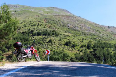   Yolun sol tarafında kırmızı bir enduro motosikleti, arka planda orman var ve asfalt zeminin üzerinde duruyor. 