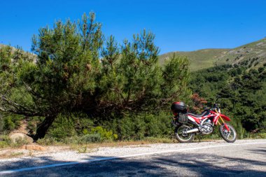   Yolun sol tarafında kırmızı bir enduro motosikleti, arka planda orman var ve asfalt zeminin üzerinde duruyor. 