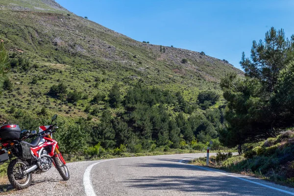   Yolun sol tarafında kırmızı bir enduro motosikleti, arka planda orman var ve asfalt zeminin üzerinde duruyor. 