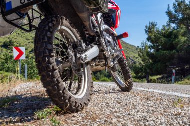 Asfalt yolun kenarındaki çakıllı yolda park edilmiş kırmızı bir enduro motosikleti. Sadece vites lastiği ve mekanik parçalar var. Çerçevede siyah toprak yolda. Lastik, motosiklet, yer manzaralı. 