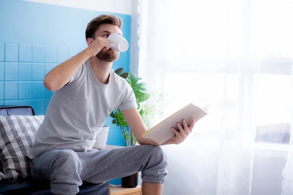 Młody człowiek czytanie książki i picie kawy na kanapie w żywej — Zdjęcie stockowe