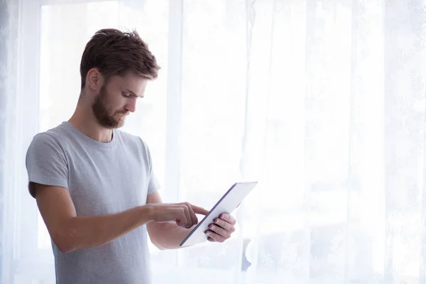 Uomo barbuto utilizzando tablet mentre in piedi vicino alla finestra nel suo m — Foto Stock