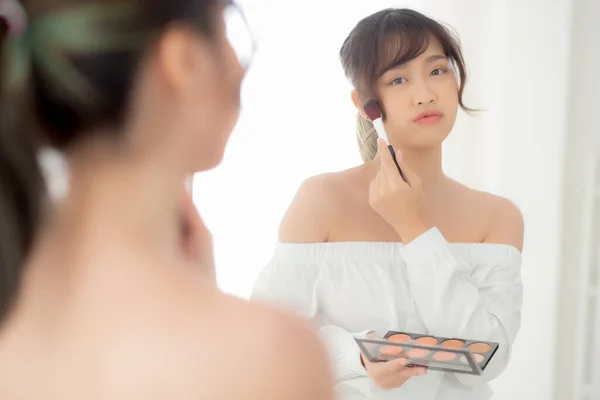 Beauty portrait young asian woman smiling with face looking mirror applying makeup with brush cheek in the bedroom, beautiful girl holding blusher, skin care and cosmetic table fashion concept.