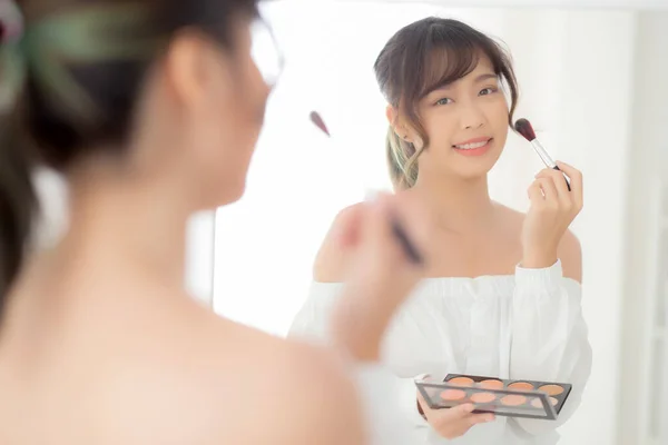 Beauty portrait young asian woman smiling with face looking mirror applying makeup with brush cheek in the bedroom, beautiful girl holding blusher, skin care and cosmetic table fashion concept.