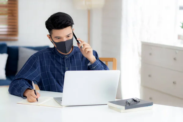 Young asian business man in face mask talking smartphone and work on laptop computer for protect covid-19, businessman quarantine work from home and writing notebook, social distancing and stay home.