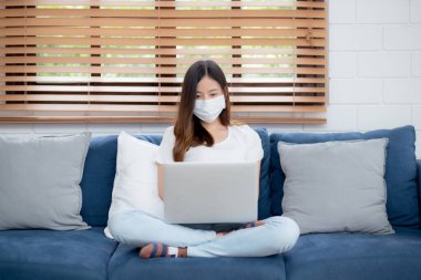 Yüz maskeli Asyalı genç bir iş kadını evde dizüstü bilgisayardan internete, oturma odasında internete, tıbbi maskeli bir kız ve covid-19 salgını için karantinaya alınıyor..